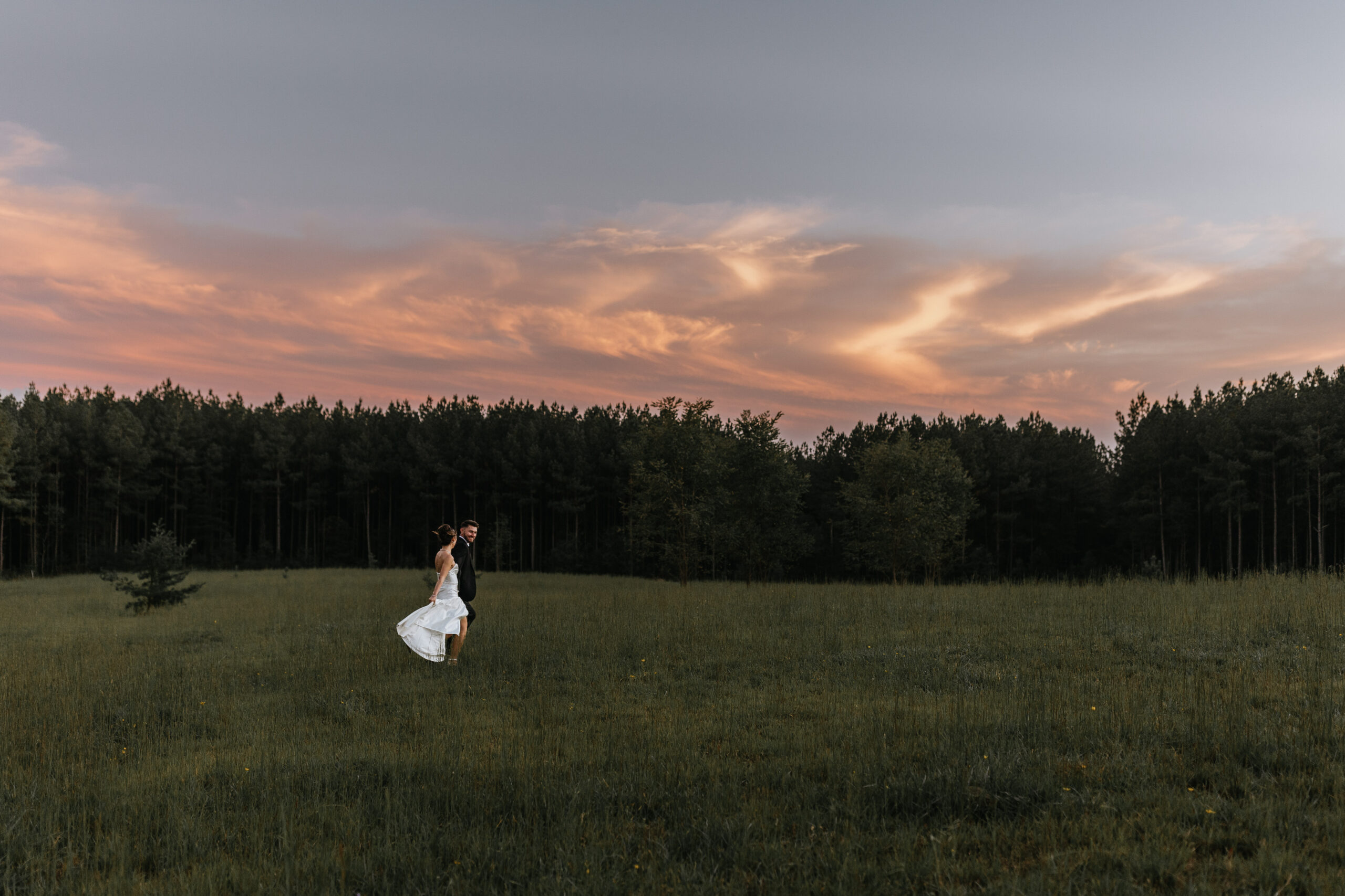 bride and groom walk away in field leading to a pine forest as the sun sets