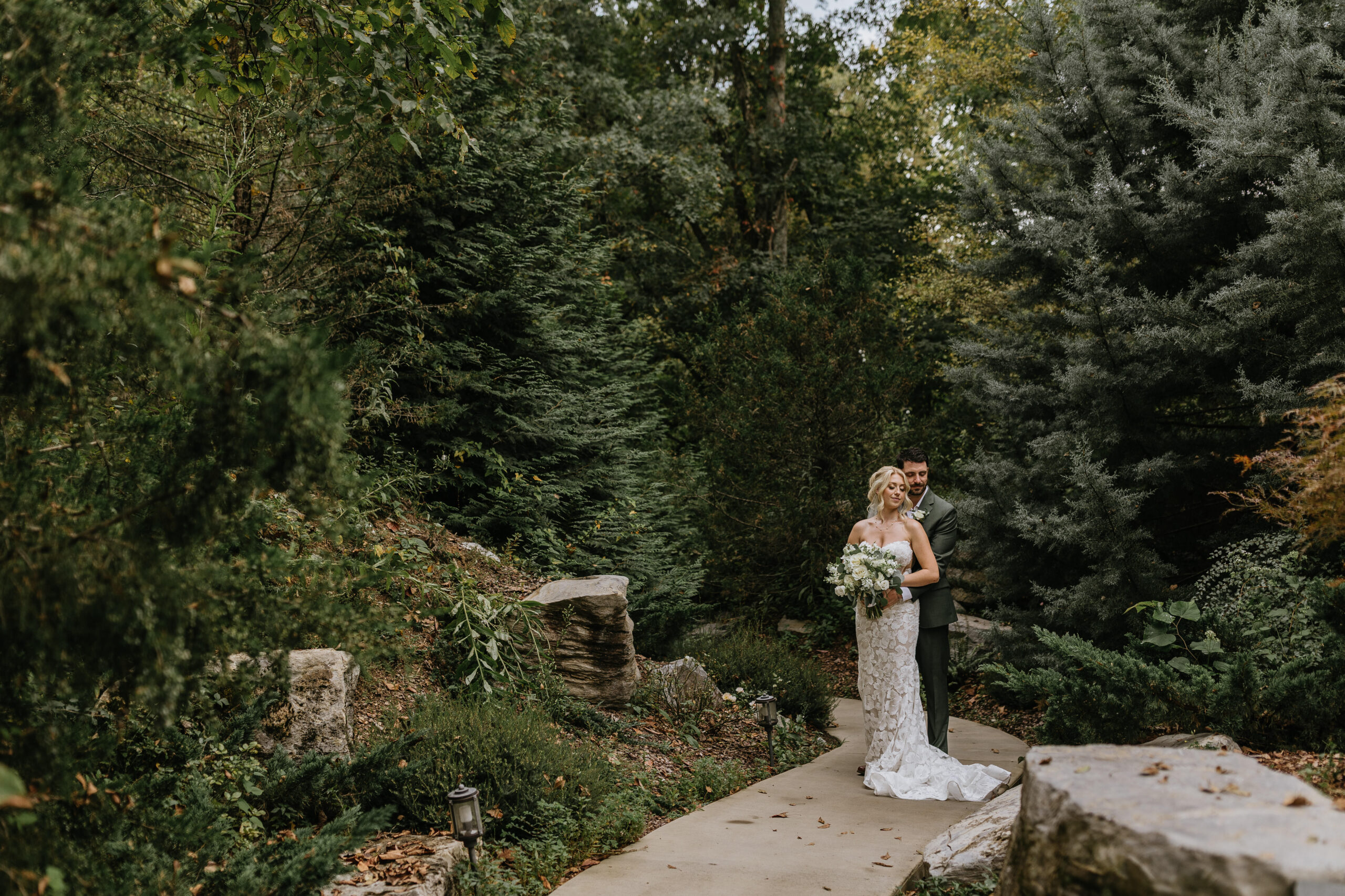 bride and groom stand in forest and and bride leans back against groom during their waterstone wedding